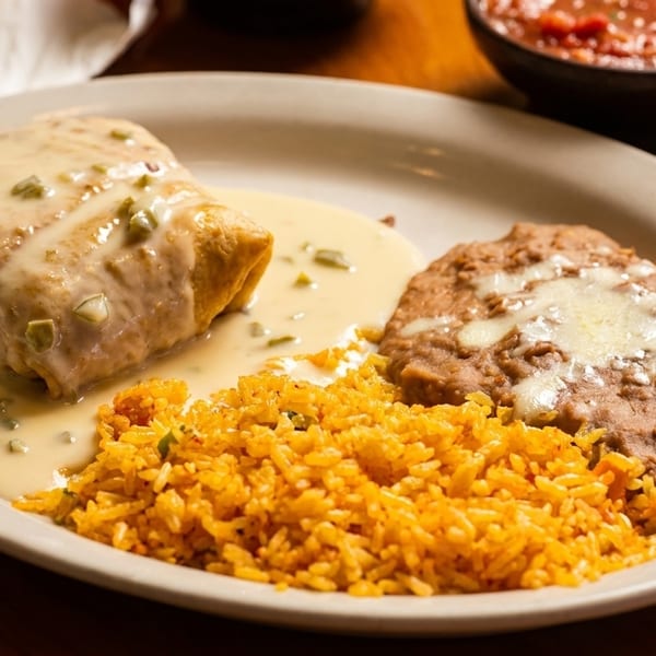 A burrito covered in white sauce with rice and refried beans on a white plate.