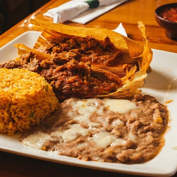 A plate of tamales, rice, and refried beans with melted cheese, alongside a side salad and salsa.