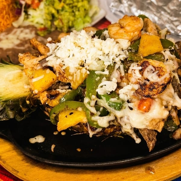 Pineapple filled with fajita mix, shrimp, peppers, and cheese, with rice and beans in the background.