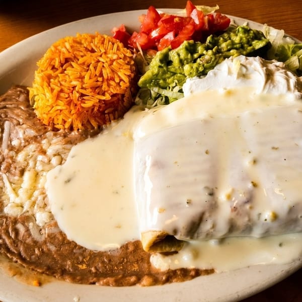A large plate of Mexican food with a burrito covered in white sauce, rice, beans, guacamole, and salsa.