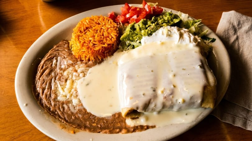 A large plate of Mexican food with a burrito covered in white sauce, rice, beans, guacamole, and salsa.